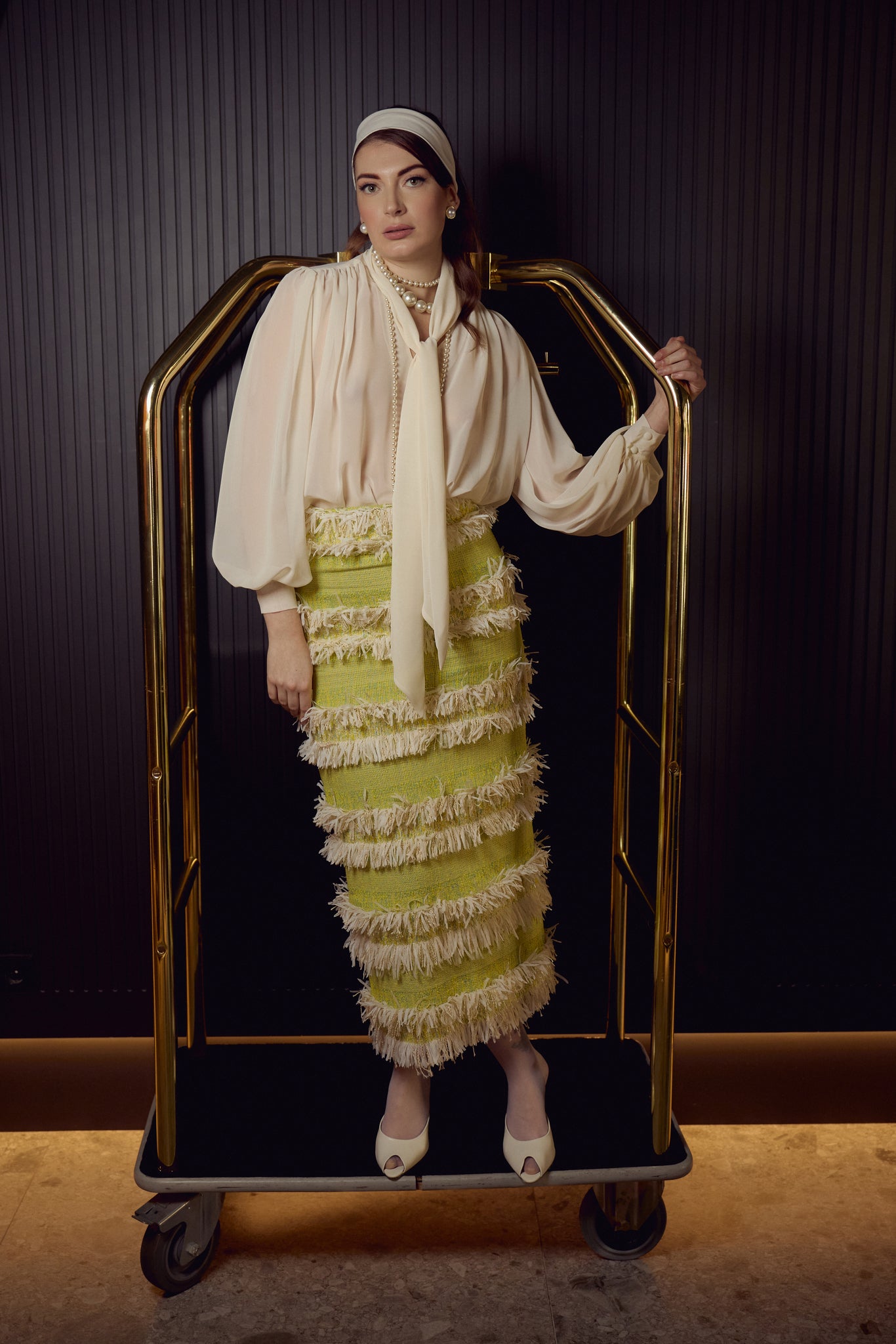 Plano general del look de invitada de la colección Suite Primavera Verano 2026 de Zinnia Brand con blusa beige de gasa de manga larga y lazada en el cuello combinada con falda midi de tweed lima con flecos beige. Un diseño elegante y sofisticado de inspiración contemporánea.
