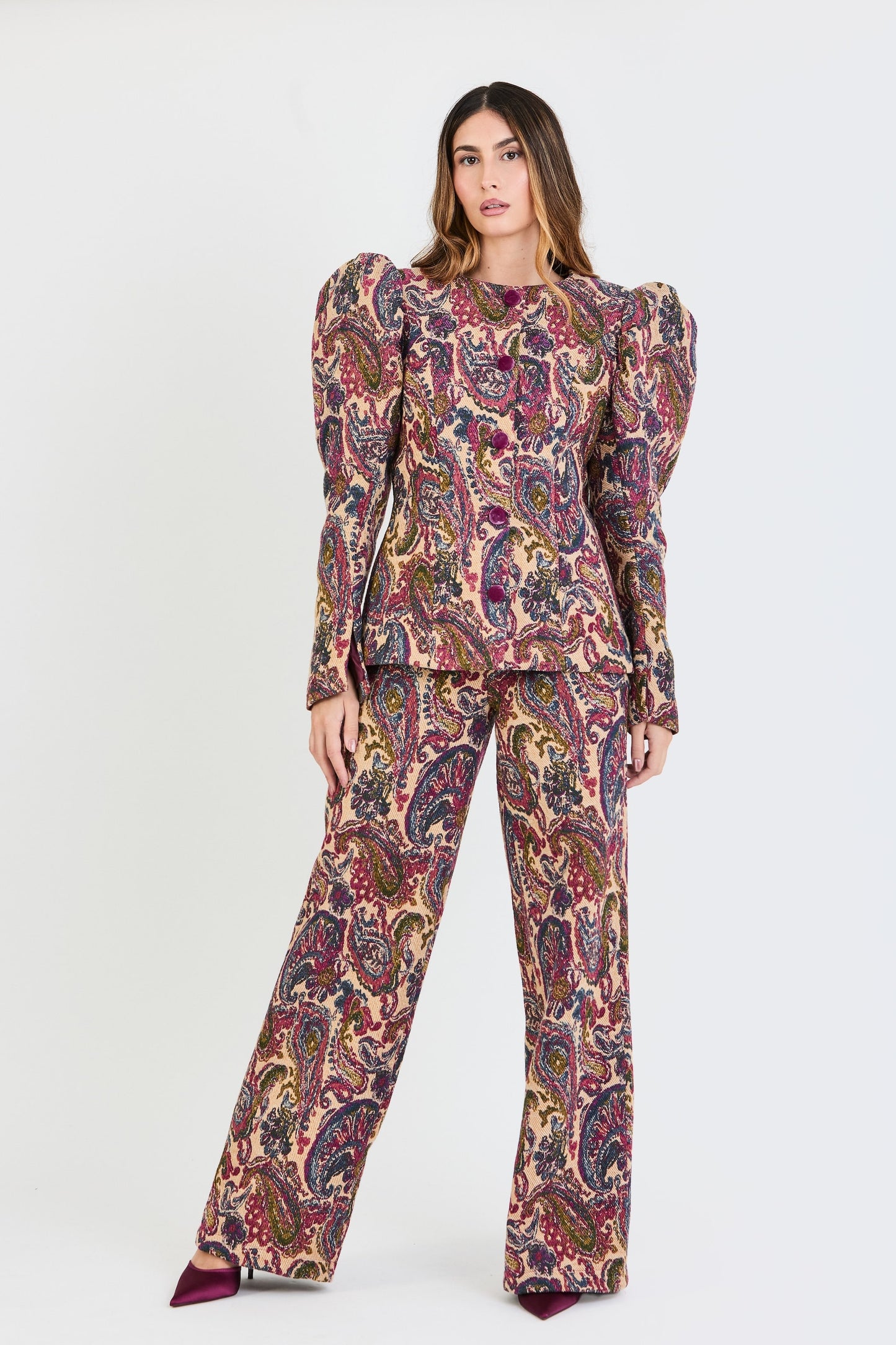 Plano general del traje de invitada de la colección Otoño Invierno 2025-2026 de Zinnia Brand con estampado cashmere en color fucsia, azul y beige. La chaqueta de cuello redondo, con botones forrados en terciopelo magenta. Las mangas son largas y abullonadas. El pantalón es del mismo estampado de corte recto. Aporta elegancia contemporánea.
