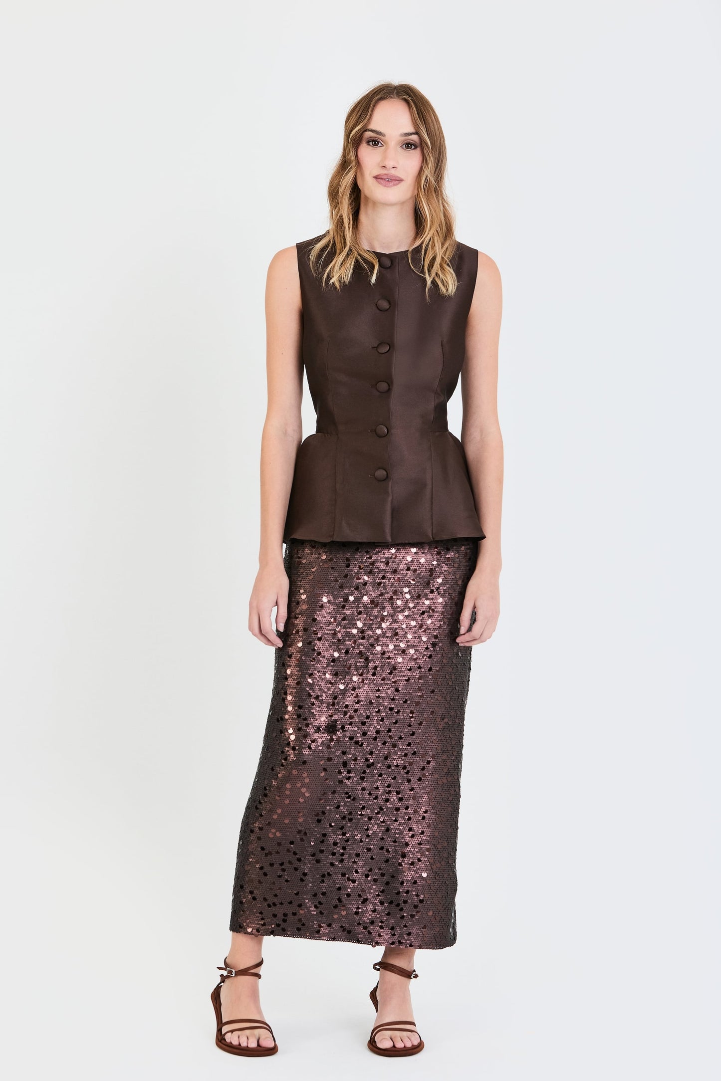 Plano general del conjunto de invitada de la colección Otoño Invierno 2025-2026 de Zinnia Brand con top estructurado en tono marrón y detalle peplum en la cintura, botones forrados  y falda de lentejuelas marrón. Un diseño elegante y contemporáneo que aporta sofisticación y feminidad.
