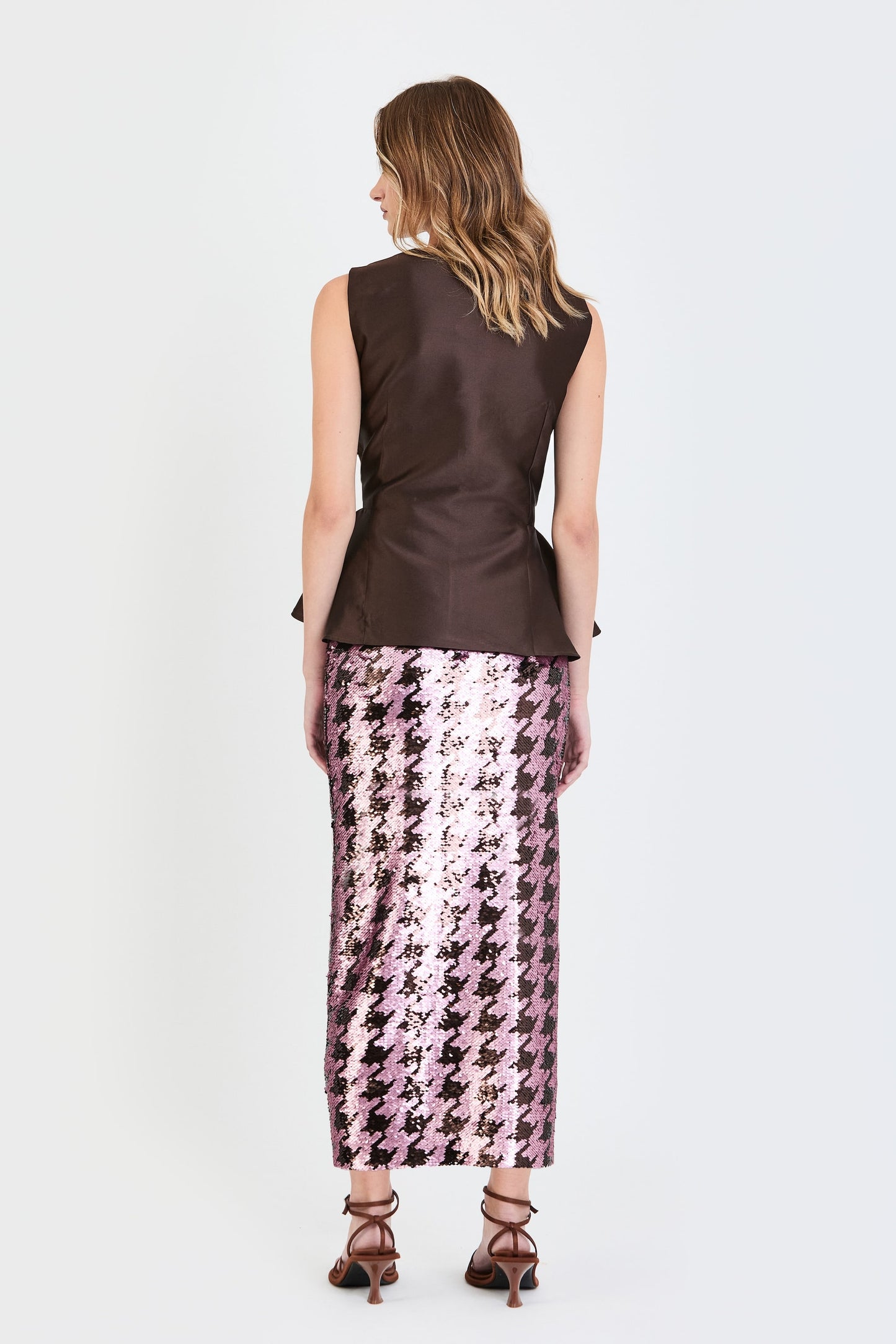 Vista de espaldas del conjunto de invitada de la colección Otoño Invierno 2025-2026 de Zinnia Brand con top estructurado en tono marrón y detalle peplum en la cintura, botones forrados  y falda de lentejuelas estampado pata de gallo ma