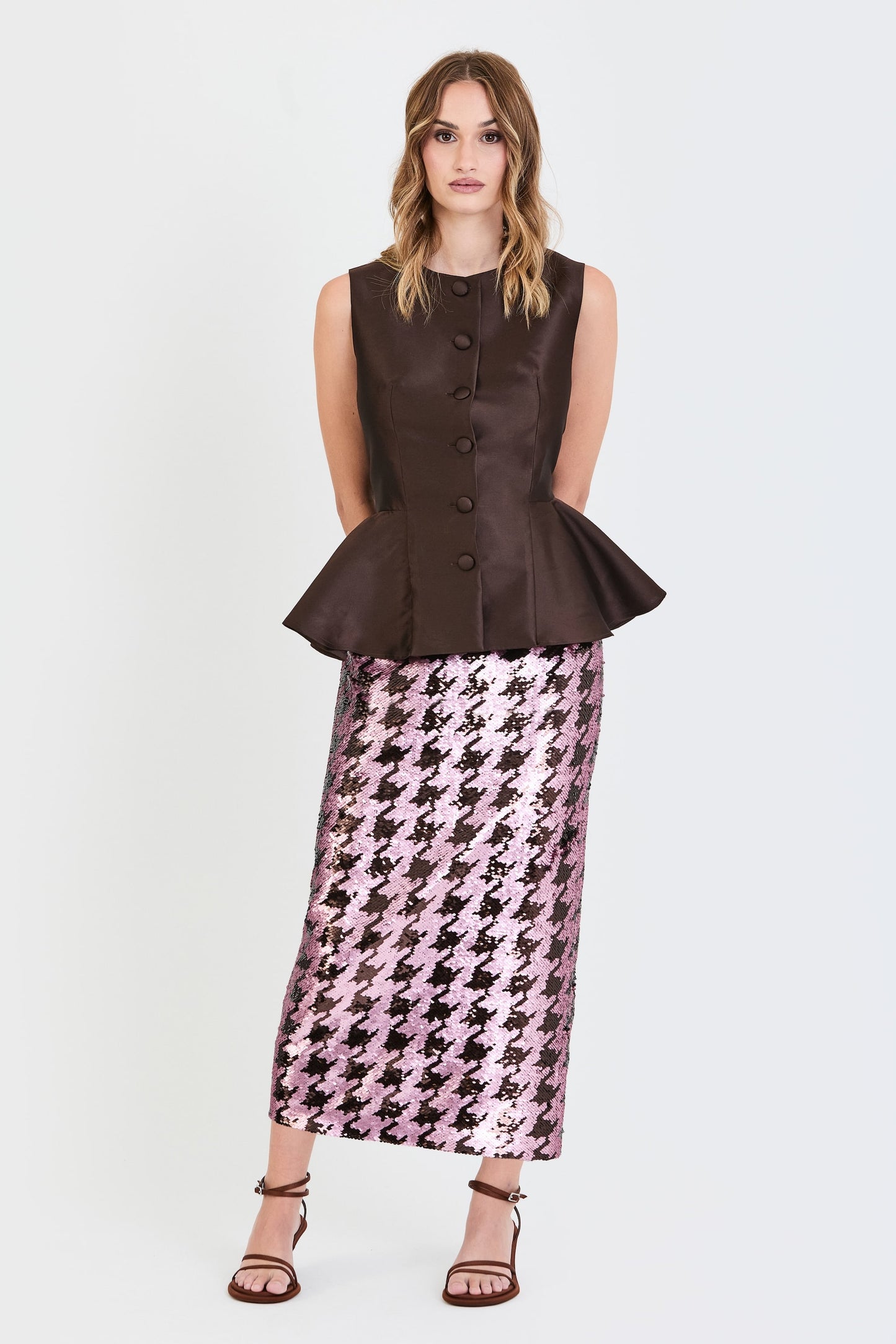Plano general del conjunto de invitada de la colección Otoño Invierno 2025-2026 de Zinnia Brand con top estructurado en tono marrón y detalle peplum en la cintura, botones forrados  y falda de lentejuelas estampado pata de gallo marrón y rosa. Un diseño elegante y contemporáneo que aporta sofisticación y feminidad.
