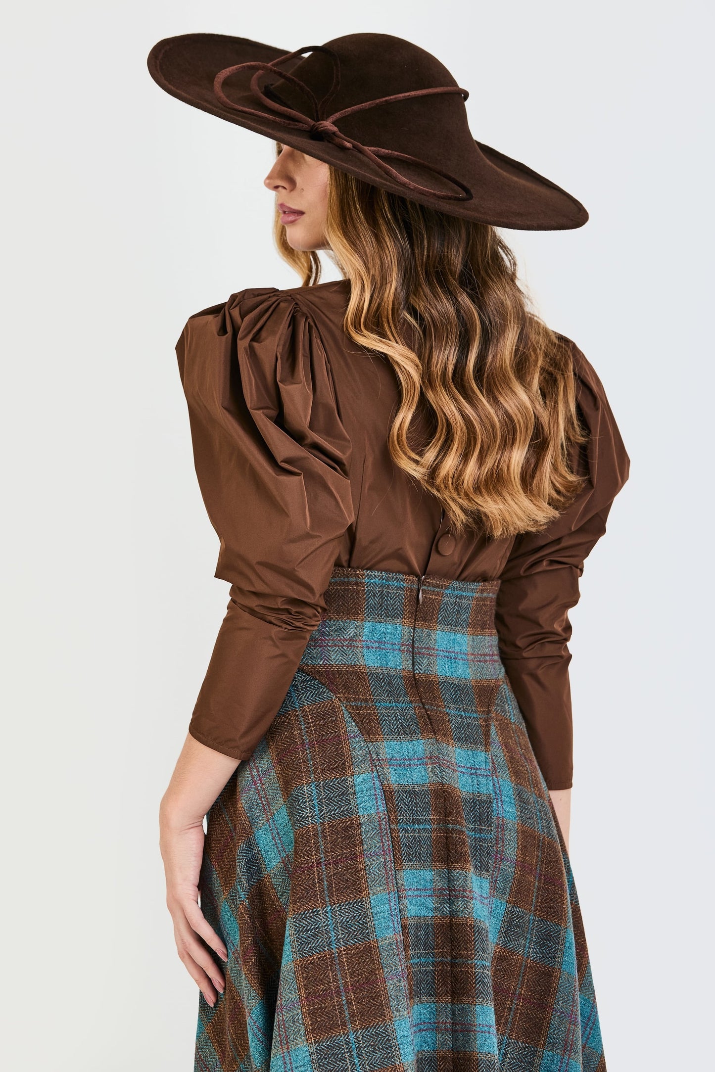 Primer plano de espalda del conjunto de invitada de la colección Otoño Invierno 2025-2026 de Zinnia Brand con blusa marrón de mangas abullonadas y falda midi de cuadros en color marrón y azul. Un diseño femenino y refinado que aporta estilo y personalidad para celebraciones.

