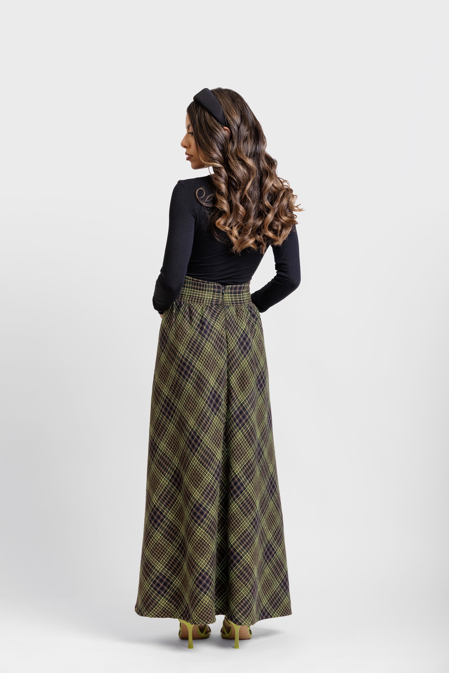 Vista de espalda de falda midi de invitada de la colección Otoño Invierno 2025-2026 de Zinnia Brand con estampado de paño de cuadros en color marino, negro y pistacho. Talle alto, realzando la figura y a la altura de la caderas sale un pequeño vuelo. Lleva una botones de terciopelo negro. Diseño elegante y contemporáneo.
