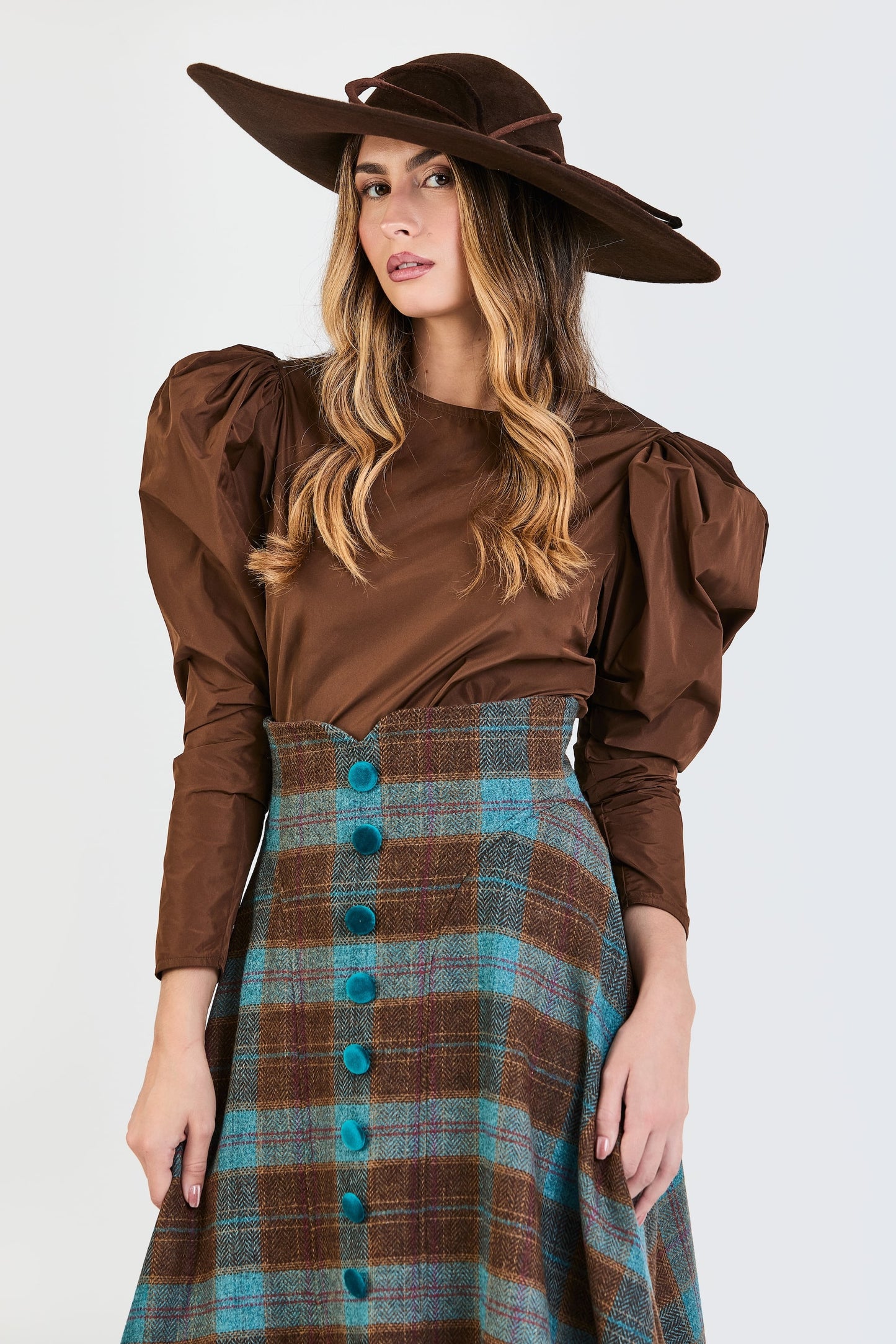 Primer plano del look de invitada de la colección Otoño Invierno 2025-2026 de Zinnia Brand con blusa marrón de mangas abullonadas y falda midi de cuadros en color marrón y azul. Un diseño que combina elegancia, textura y sofisticación contemporánea.
