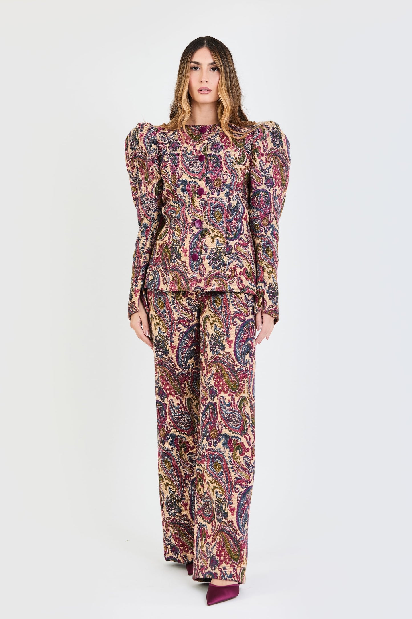 Plano general del traje de invitada de la colección Otoño Invierno 2025-2026 de Zinnia Brand con estampado cashmere en color fucsia, azul y beige. La chaqueta de cuello redondo, con botones forrados en terciopelo magenta. Las mangas son largas y abullonadas. El pantalón es del mismo estampado de corte recto. Aporta elegancia contemporánea.
